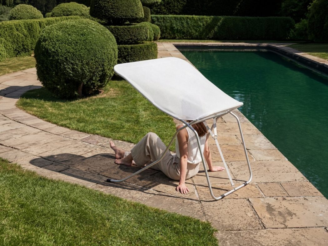 Person sits on a stone poolside, shaded by a white canopy chair, by a green hedge and clear pool water.