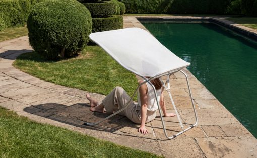 Person sits on a stone poolside, shaded by a white canopy chair, by a green hedge and clear pool water.