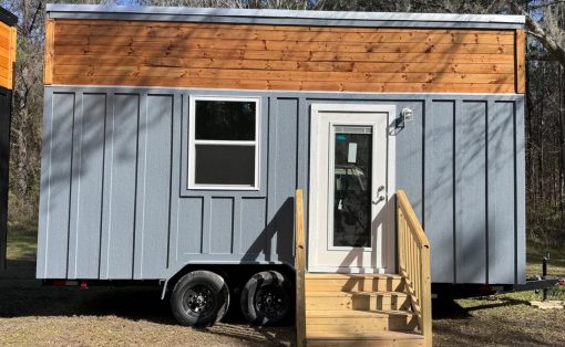 Blue-gray tiny house on a dual-axle trailer with a wooden upper facade, white-framed window, and a wooden staircase leading to a glass-panel front door.