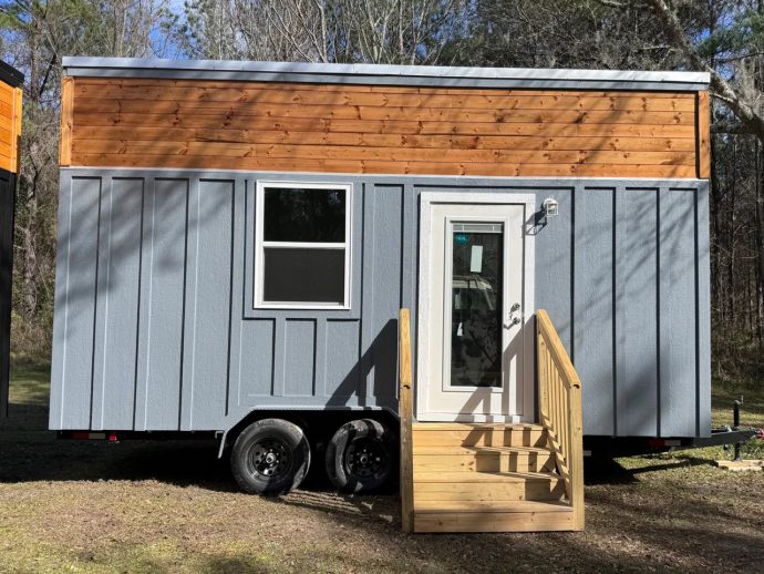 Blue-gray tiny house on a dual-axle trailer with a wooden upper facade, white-framed window, and a wooden staircase leading to a glass-panel front door.