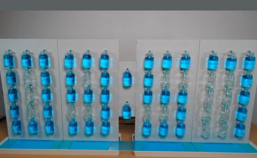 Indoor science exhibit featuring two white panels with vertical rows of blue liquid-filled bottles connected by tubing, forming a cascading display on a wooden table base.