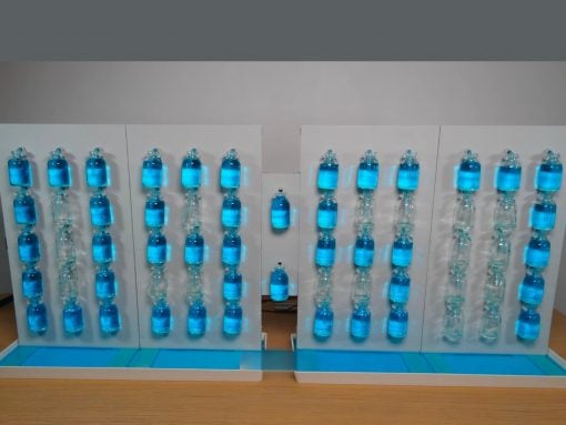Indoor science exhibit featuring two white panels with vertical rows of blue liquid-filled bottles connected by tubing, forming a cascading display on a wooden table base.