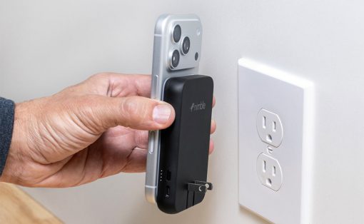 Hand holding a smartphone with a black portable power bank attached, as the plug is inserted into a wall outlet nearby, ready to charge the phone.