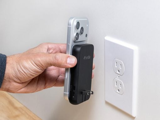Hand holding a smartphone with a black portable power bank attached, as the plug is inserted into a wall outlet nearby, ready to charge the phone.