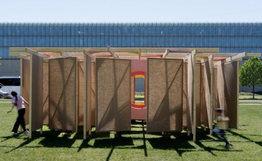 Outdoor wooden panel maze sculpture on a grassy field, pink circular center with orange accents; a person and a child move nearby.