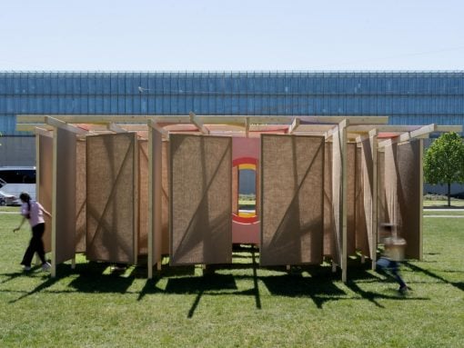 Outdoor wooden panel maze sculpture on a grassy field, pink circular center with orange accents; a person and a child move nearby.