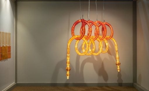 Glowing orange and yellow looped sculpture hanging from the ceiling in an art gallery, forming intertwined rings.
