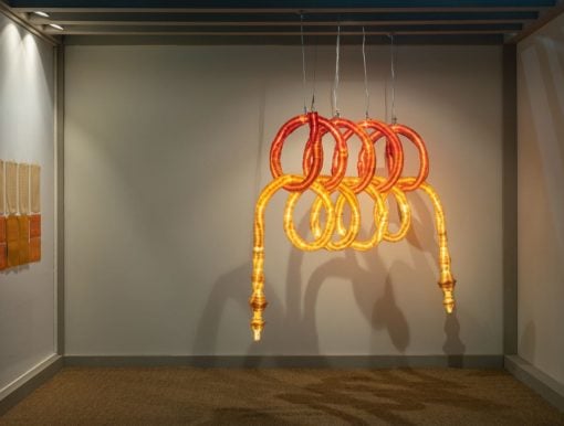 Glowing orange and yellow looped sculpture hanging from the ceiling in an art gallery, forming intertwined rings.
