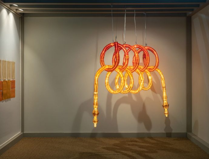 Glowing orange and yellow looped sculpture hanging from the ceiling in an art gallery, forming intertwined rings.