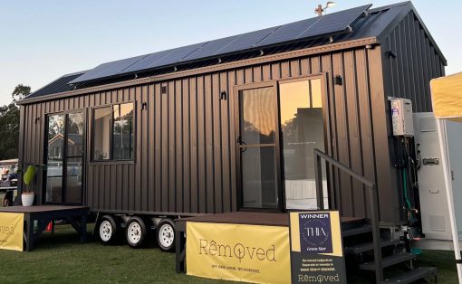 Dark gray tiny house on wheels with solar panels on the roof, glass doors, and steps, parked on a grassy field with yellow banners nearby.