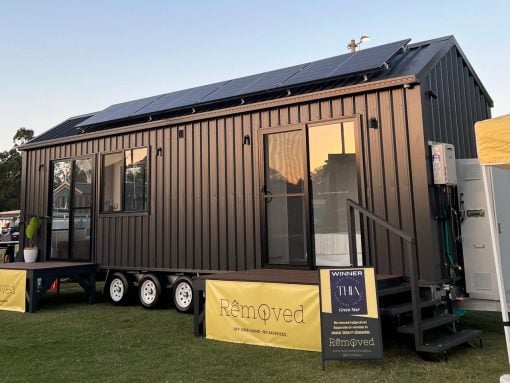Dark gray tiny house on wheels with solar panels on the roof, glass doors, and steps, parked on a grassy field with yellow banners nearby.