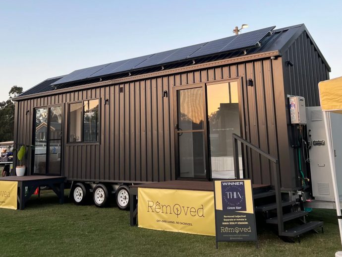 Dark gray tiny house on wheels with solar panels on the roof, glass doors, and steps, parked on a grassy field with yellow banners nearby.