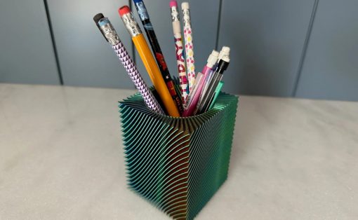 Green accordion-folded pencil cup holding assorted pencils and pens on a light desk surface behind it a gray wall/cabinet.