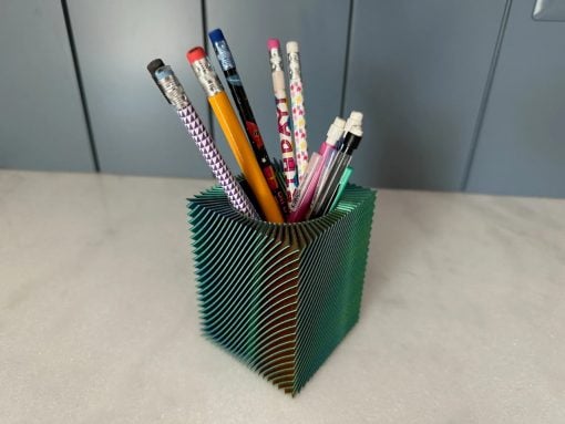Green accordion-folded pencil cup holding assorted pencils and pens on a light desk surface behind it a gray wall/cabinet.