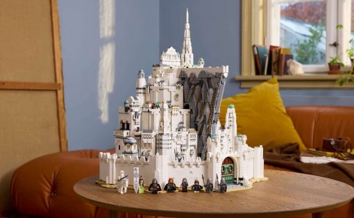 Large white LEGO fortress with towers built on a rocky cliff, set on a wooden table indoors, with minifigures lined up at the front.
