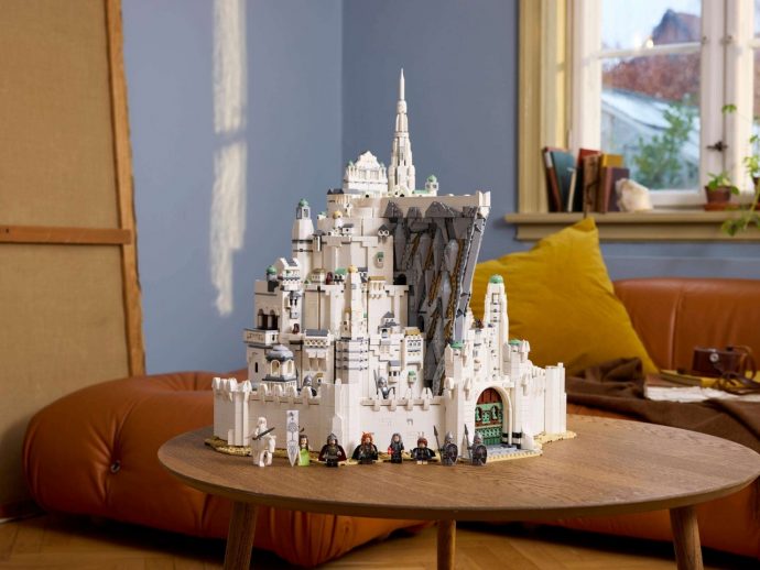 Large white LEGO fortress with towers built on a rocky cliff, set on a wooden table indoors, with minifigures lined up at the front.