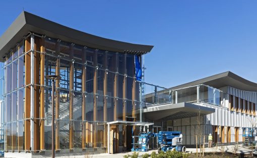 Modern glass-and-wood building under construction with scaffolding and a blue sky backdrop.