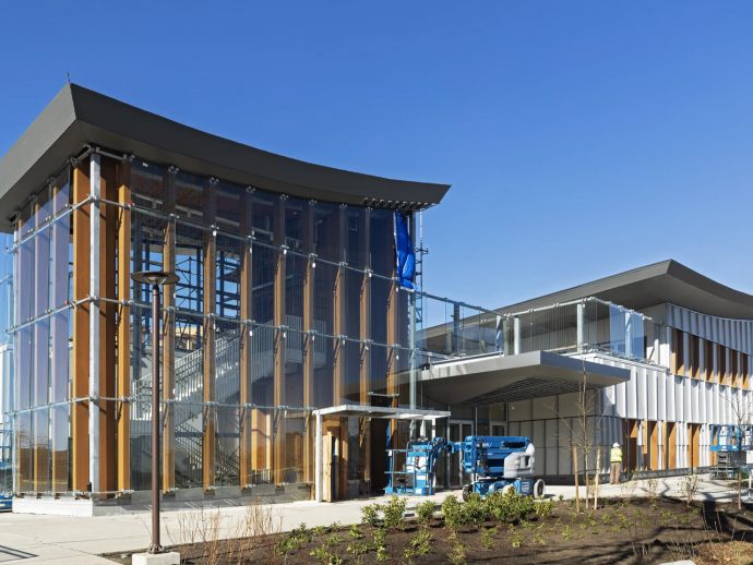 Modern glass-and-wood building under construction with scaffolding and a blue sky backdrop.