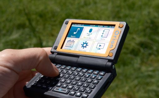 Handheld portable device with a fold-out keyboard and colorful touchscreen icons, held over green grass outdoors.