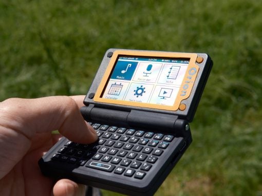 Handheld portable device with a fold-out keyboard and colorful touchscreen icons, held over green grass outdoors.