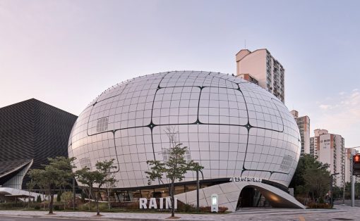 Futuristic white dome building with a grid pattern on a city street, with trees in front and the word 'RAINI' on the facade.