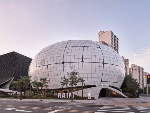 Futuristic white dome building with a grid pattern on a city street, with trees in front and the word 'RAINI' on the facade.