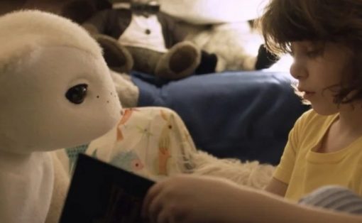 Child in a yellow shirt reading a book to a large white stuffed bear on a bed. (Informative scene)