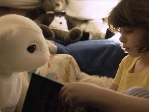 Child in a yellow shirt reading a book to a large white stuffed bear on a bed. (Informative scene)