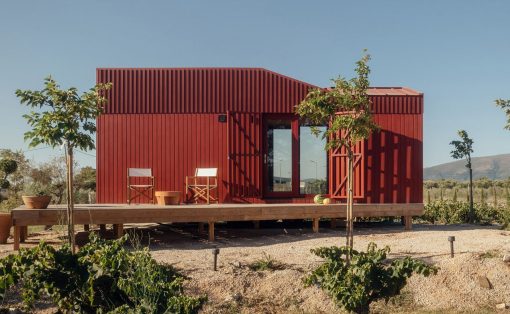 Red corrugated cabin on a raised wooden deck with two chairs and a glass door, set in a sunny rural landscape.