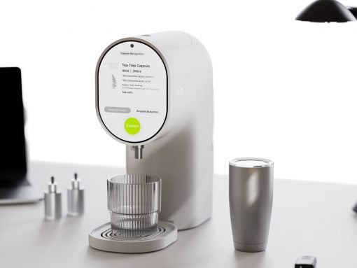 White, modern coffee machine with a circular touchscreen, brewing into a glass cup on a white desk.