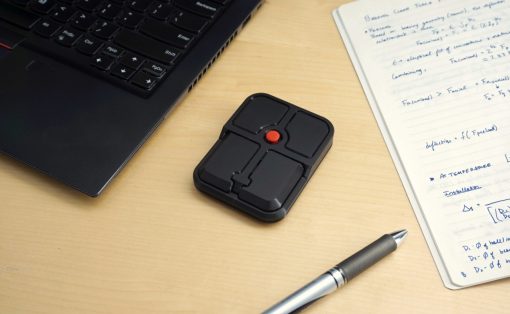 Small black handheld multi-button input device with four square pads and a red center button on a wooden desk beside a laptop and notebook.