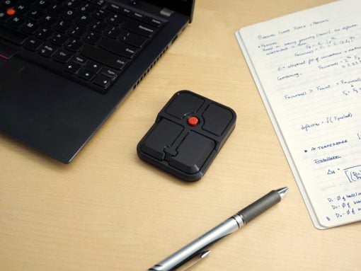 Small black handheld multi-button input device with four square pads and a red center button on a wooden desk beside a laptop and notebook.