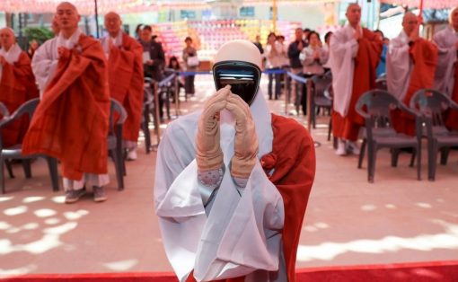 A monk in white robes with a futuristic visor on the face holds a prayer gesture as orange-robed monks stand in the background under colorful lanterns at a ceremonial event.