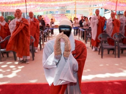 A monk in white robes with a futuristic visor on the face holds a prayer gesture as orange-robed monks stand in the background under colorful lanterns at a ceremonial event.