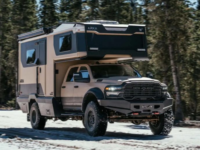 Beige heavy-duty pickup with a large rooftop camper extending over the cab, driving on a snowy road with trees in the background.