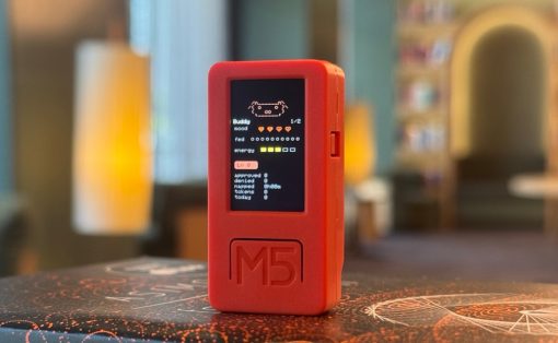 Red handheld M5 device with a small black screen showing pixel art, resting on a patterned table in a cozy room.