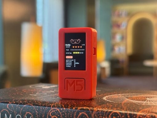 Red handheld M5 device with a small black screen showing pixel art, resting on a patterned table in a cozy room.