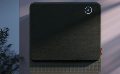 Square black fabric-covered wall device with rounded corners, circular status light in the top-right, and perforated base; likely a smart air purifier or speaker with a camera nearby.