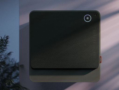 Square black fabric-covered wall device with rounded corners, circular status light in the top-right, and perforated base; likely a smart air purifier or speaker with a camera nearby.