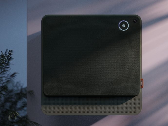 Square black fabric-covered wall device with rounded corners, circular status light in the top-right, and perforated base; likely a smart air purifier or speaker with a camera nearby.