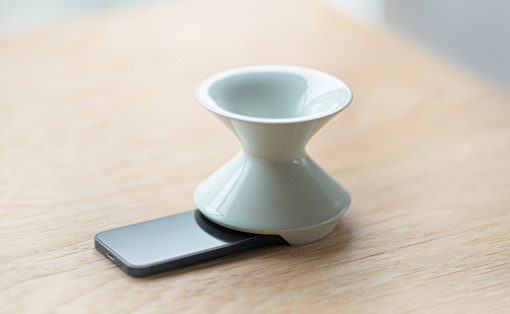 White ceramic hourglass-shaped object resting on a smartphone lying on a wooden table; appears to be a small decorative or functional stand or holder atop the device.