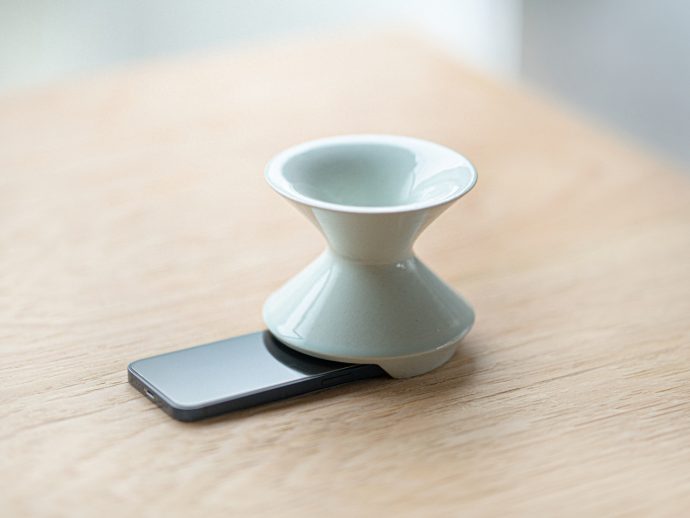 White ceramic hourglass-shaped object resting on a smartphone lying on a wooden table; appears to be a small decorative or functional stand or holder atop the device.