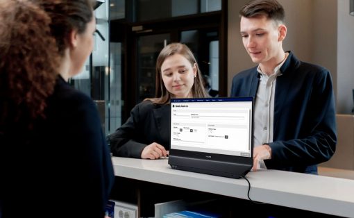 Two front-desk staff assist a guest at a counter with a laptop displaying a hotel check-in form. .