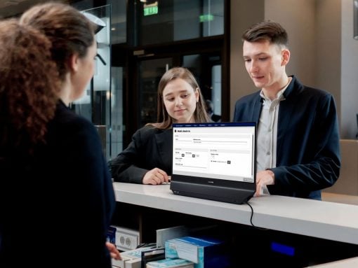 Two front-desk staff assist a guest at a counter with a laptop displaying a hotel check-in form. .