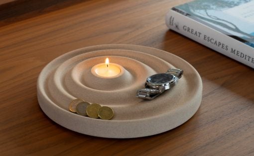 Beige concentric candle holder with a lit tealight, a silver wristwatch, and coins on a wooden table beside a travel book titled 'Great Escapes Mediterranean'.