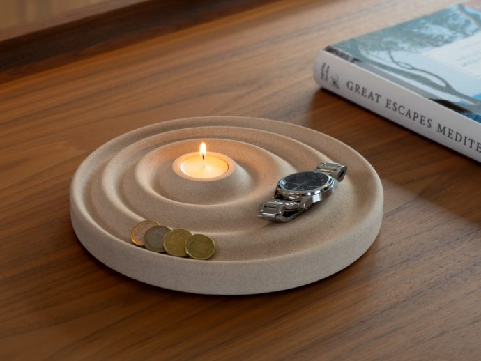 Beige concentric candle holder with a lit tealight, a silver wristwatch, and coins on a wooden table beside a travel book titled 'Great Escapes Mediterranean'.