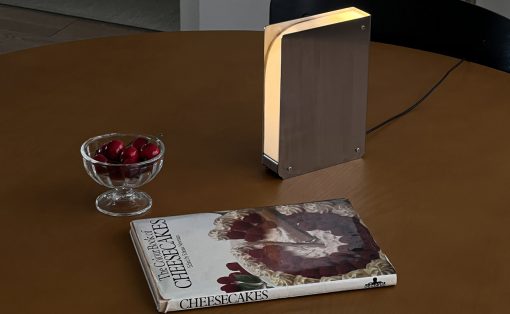 Rectangular metal lamp glows on a round wooden table beside a bowl of cherries and a cheesecake cookbook.