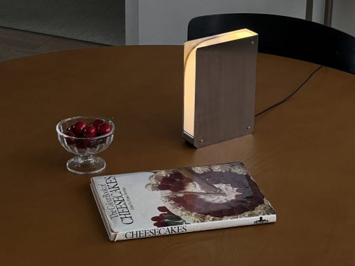 Rectangular metal lamp glows on a round wooden table beside a bowl of cherries and a cheesecake cookbook.