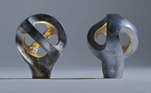 Two weathered metal sculptures with circular cutouts stand on a gray surface in a neutral studio, gold inner rings visible through the holes.