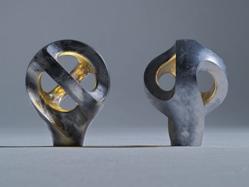 Two weathered metal sculptures with circular cutouts stand on a gray surface in a neutral studio, gold inner rings visible through the holes.